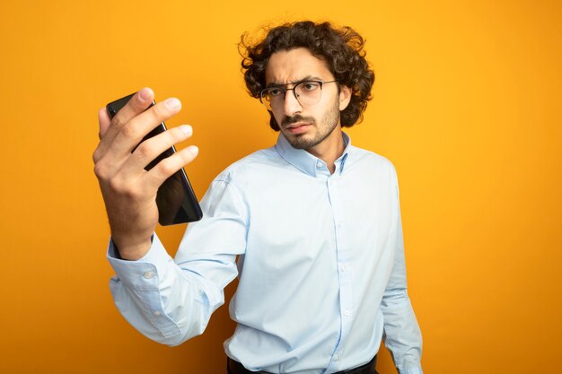 Fronçant les sourcils jeune bel homme caucasien portant des lunettes tenant et regardant téléphone mobile isolé sur fond orange avec espace de copie