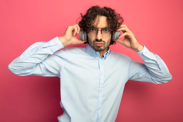 Fronçant les sourcils jeune bel homme portant des lunettes et des écouteurs en regardant les écouteurs de saisie avant isolé sur mur rose