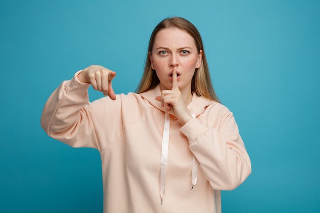 Fronçant les sourcils jeune femme blonde regardant et pointant vers la caméra faisant le geste de silence