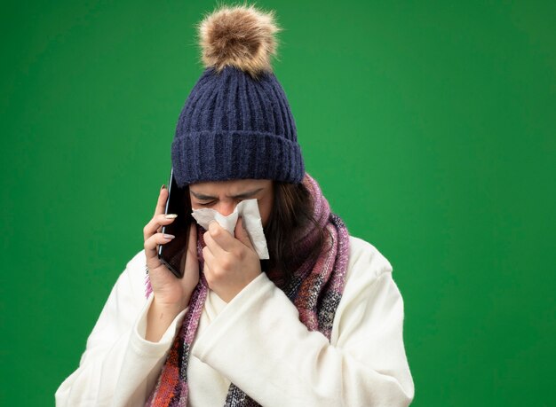 Fronçant les sourcils jeune femme malade portant chapeau d'hiver robe et écharpe parler au téléphone essuyant le nez avec serviette avec les yeux fermés isolé sur mur vert