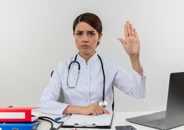 Photo gratuite fronçant les sourcils jeune femme médecin portant une robe médicale et un stéthoscope assis au bureau avec des outils médicaux et un ordinateur portable faisant le geste d'arrêt isolé sur un mur blanc