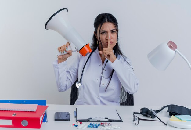 Fronçant les sourcils jeune femme médecin portant une robe médicale et un stéthoscope assis au bureau avec des outils médicaux tenant le haut-parleur à la recherche de faire silence geste isolé