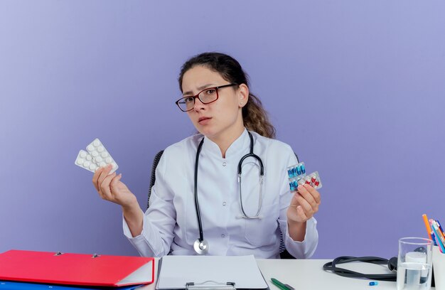Fronçant les sourcils jeune femme médecin portant une robe médicale et un stéthoscope assis au bureau avec des outils médicaux tenant des pilules médicales à la recherche d'isolement