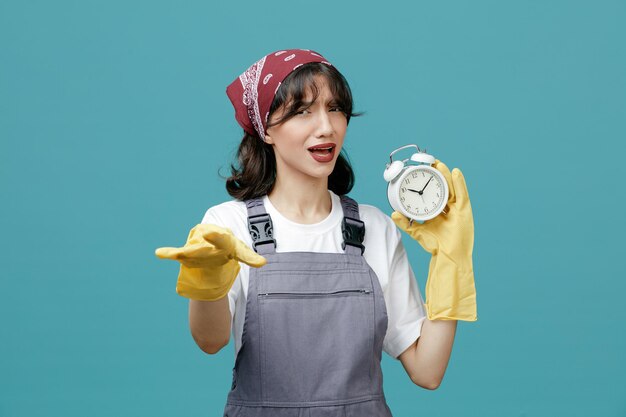 Fronçant les sourcils jeune femme nettoyeuse portant un bandana uniforme et des gants en caoutchouc montrant un réveil regardant la caméra montrant un geste de départ isolé sur fond bleu