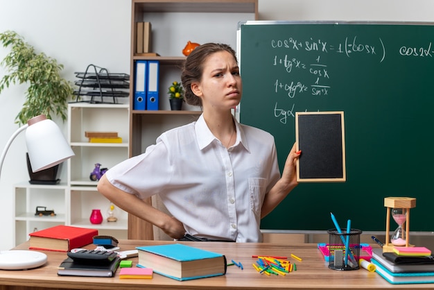 Fronçant les sourcils jeune femme professeur de mathématiques assis au bureau avec des fournitures scolaires tenant un mini tableau noir gardant la main sur la taille regardant à l'avant en classe