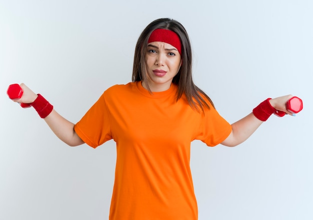 Photo gratuite fronçant les sourcils jeune femme sportive portant un bandeau et des bracelets étirant des haltères isolés sur un mur blanc