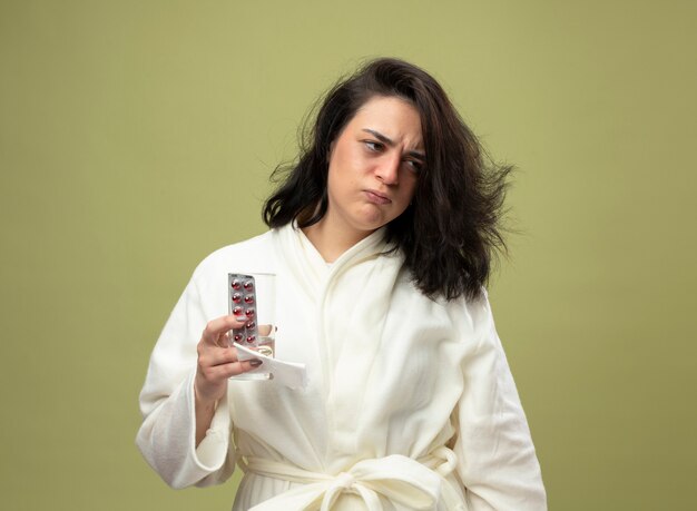 Fronçant les sourcils jeune fille malade de race blanche portant robe holding pack de pilules médicales verre d'eau et serviette à côté isolé sur fond vert olive