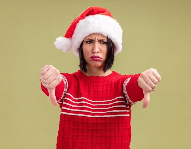 Fronçant les sourcils jeune fille portant bonnet de Noel à côté montrant les pouces vers le bas isolé sur fond vert olive