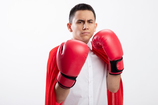 Fronçant les sourcils jeune garçon de super-héros en cape rouge portant des gants de boîte regardant la caméra faisant le geste de boxe isolé sur fond blanc avec espace de copie