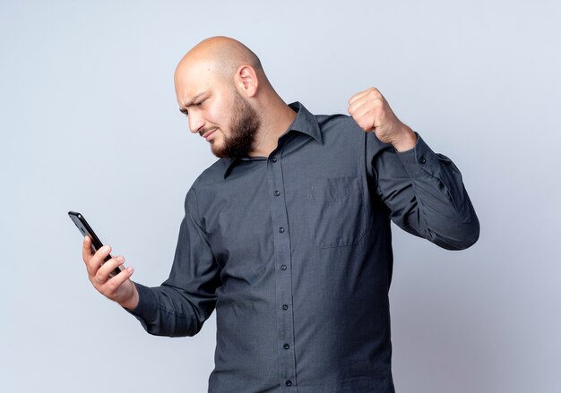 Fronçant les sourcils jeune homme de centre d'appels chauve tenant et regardant le téléphone mobile et levant le poing isolé sur blanc