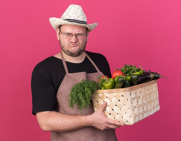Photo gratuite fronçant les sourcils jeune homme jardinier portant un chapeau de jardinage tenant un panier de légumes isolé sur un mur rose
