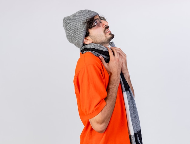 Fronçant les sourcils jeune homme malade caucasien portant des lunettes chapeau d'hiver et écharpe debout en vue de profil mettant les mains sur l'écharpe en levant isolé sur fond blanc avec espace copie