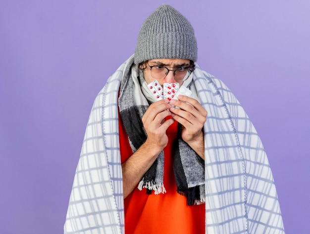 Photo gratuite fronçant les sourcils jeune homme malade portant des lunettes chapeau d'hiver et une écharpe enveloppée dans un plaid tenant des paquets de pilules médicales devant la bouche en les regardant isolés sur mur violet