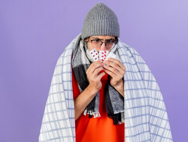 Fronçant les sourcils jeune homme malade portant des lunettes chapeau d'hiver et une écharpe enveloppée dans un plaid tenant des paquets de pilules médicales devant la bouche en les regardant isolés sur mur violet