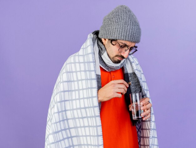 Fronçant les sourcils jeune homme malade portant des lunettes chapeau d'hiver et une écharpe enveloppée dans un plaid verser le médicament en verre dans un verre d'eau isolé sur mur violet