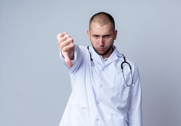 Fronçant les sourcils jeune homme médecin portant une robe médicale et un stéthoscope autour de son cou montrant le pouce vers le bas isolé sur blanc avec copie espace
