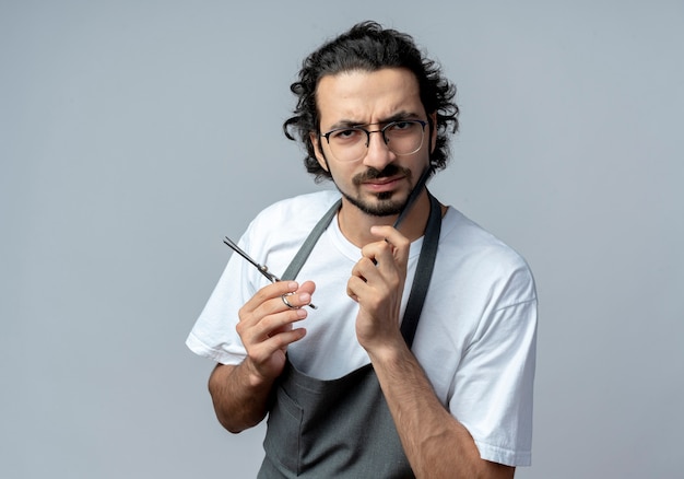 Fronçant les sourcils jeune homme de race blanche coiffeur portant des lunettes et bande de cheveux ondulés en uniforme tenant des ciseaux et toucher le visage avec un peigne isolé sur fond blanc avec espace de copie