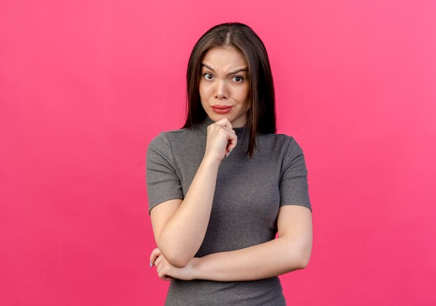 Fronçant les sourcils jeune jolie femme mettant les mains sous le coude et sur le menton isolé sur fond rose avec espace copie