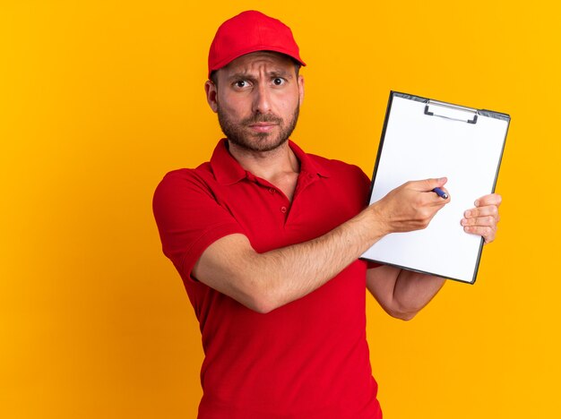 Fronçant les sourcils jeune livreur caucasien en uniforme rouge et casquette montrant le stylo pointant le presse-papiers dessus