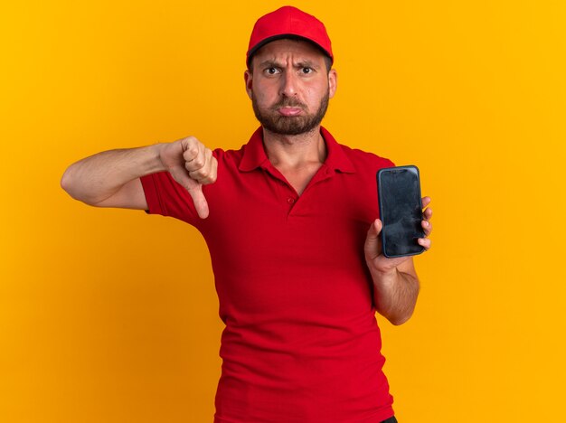 Fronçant les sourcils jeune livreur caucasien en uniforme rouge et casquette montrant un téléphone portable et le pouce vers le bas