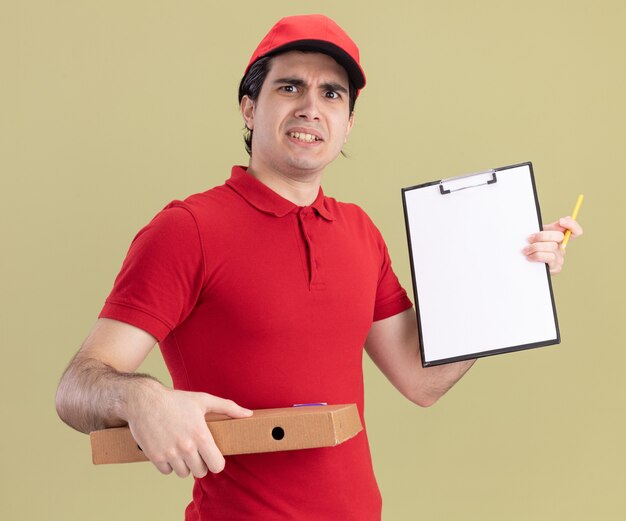 Fronçant les sourcils jeune livreur caucasien en uniforme rouge et casquette tenant un paquet de pizza montrant le presse-papiers avec un crayon à la main