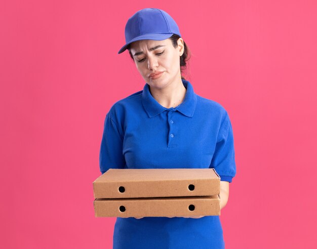 Fronçant les sourcils jeune livreuse en uniforme et casquette tenant et regardant les paquets de pizza