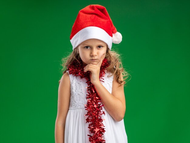 Fronçant les sourcils petite fille portant un chapeau de Noël avec guirlande sur le cou mettant la main sur le menton isolé sur mur vert