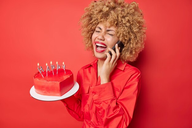 Frustrée jeune femme aux cheveux bouclés a une conversation téléphonique crie de désespoir porte une chemise tient un gâteau au coeur avec des bougies allumées bouleversé pour célébrer l'anniversaire seul isolé sur fond rouge