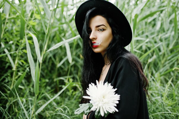 Fumeuse sensuelle fille tout en lèvres rouges noires et chapeau Goth femme dramatique tenir une fleur de chrysanthème blanche et fumer sur le roseau commun