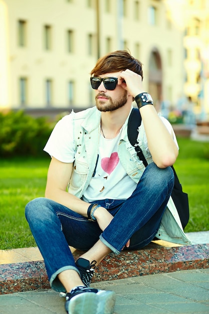 Funny smiling hipster bel homme beau en tissu d'été élégant dans la rue assis sur l'herbe dans le parc