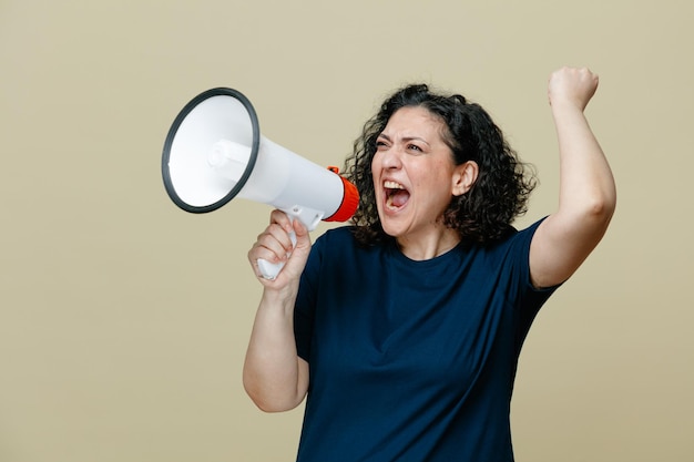 Furieuse femme d'âge moyen portant un t-shirt regardant le côté criant fort dans le haut-parleur tout en levant le poing isolé sur fond vert olive