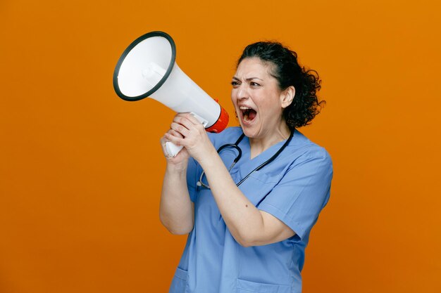 Furieuse femme médecin d'âge moyen portant l'uniforme et le stéthoscope autour du cou debout dans la vue de profil criant fort dans le haut-parleur le regardant isolé sur fond orange