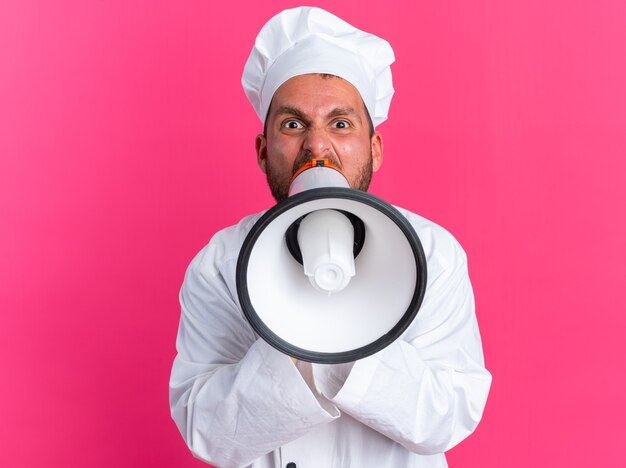 Furieux jeune cuisinier caucasien en uniforme de chef et casquette regardant la caméra criant dans un haut-parleur isolé sur un mur rose