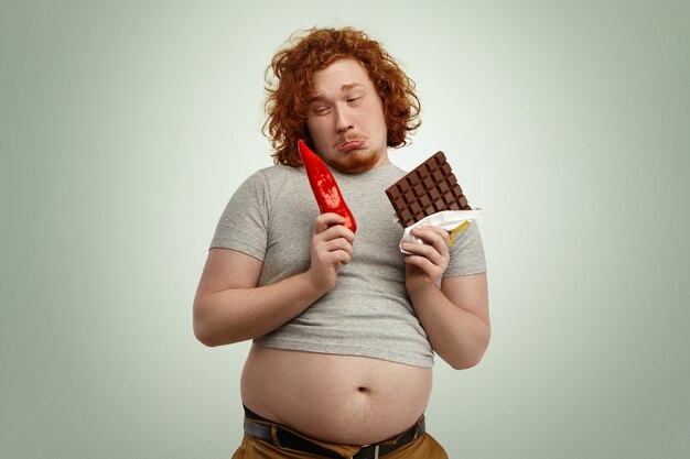 Fustré drôle jeune homme rousse portant un petit t-shirt gris avec son gros ventre qui sort, tenant du poivron rouge dans une main et une barre de chocolat dans l'autre ne veut pas manger de légumes peu appétissants