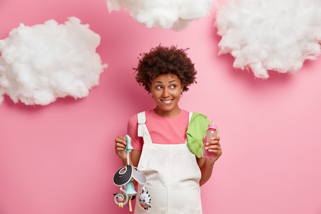 Futur concept de maternité et de grossesse. Heureuse femme enceinte tient bébé mobile et biberon, se prépare à devenir maman, attend le nouveau-né, isolé sur mur rose, nuages au-dessus