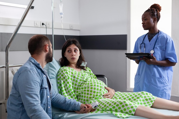 Photo gratuite les futurs parents discutent avec un assistant médical en salle d'hôpital, une femme enceinte allongée dans son lit ayant des contractions. infirmière afro-américaine préparant une patiente enceinte à une intervention chirurgicale