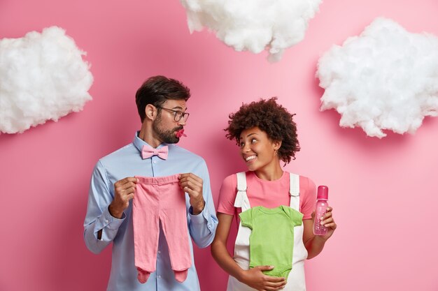 Les futurs parents heureux essaient de deviner le sexe du bébé, posent avec des curseurs pour enfants, un maillot, un biberon et une tétine, s'attendent à la naissance d'un enfant, posent contre un mur rose avec des nuages blancs moelleux au-dessus