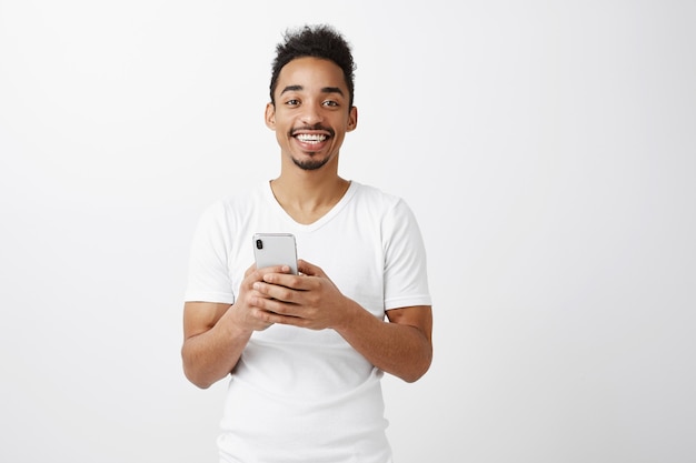 Gai attrayant mec afro-américain en t-shirt blanc tenant un téléphone mobile, souriant