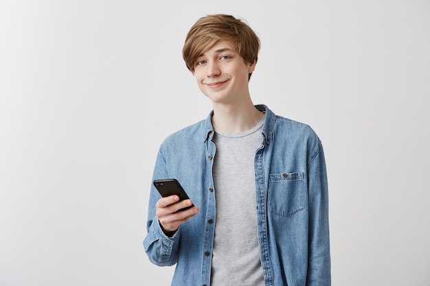 Gai étudiant blond caucasien en chemise danim naviguant sur Internet sur smartphone, se reposer après les cours, regarder l'écran et sourire. Technologies modernes et communication