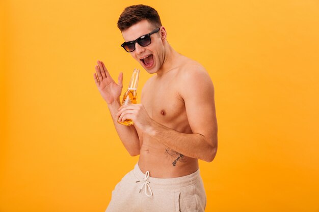 Gai homme nu en short et lunettes de soleil tenant une bouteille de bière et hurlant