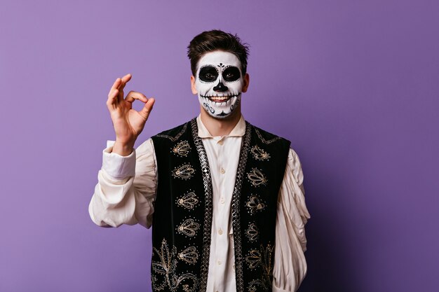 Gai mec zombie souriant sur le mur violet. Heureux jeune homme avec un maquillage effrayant posant à halloween avec signe correct.