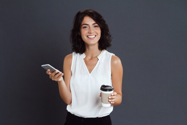 Gaie jeune femme tenant son téléphone et son café dans les mains