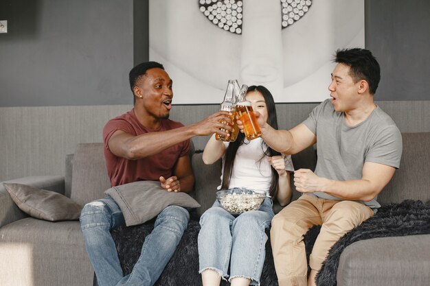 Un garçon africain et un couple asiatique tintent une bouteille avec une bière Des amis regardent un match de football en mangeant du pop-corn