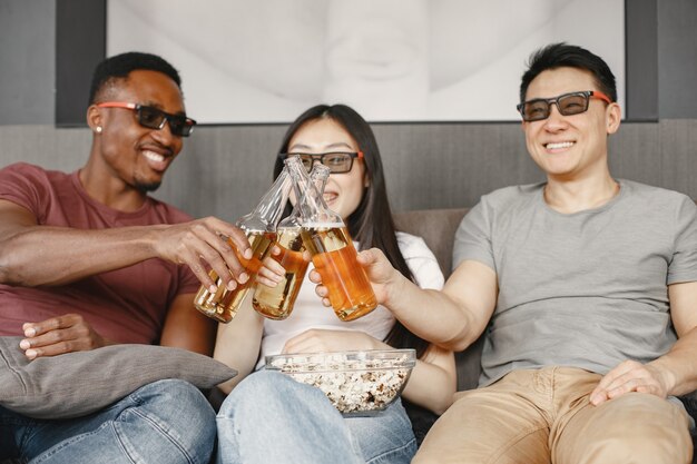 Un garçon africain et un couple asiatique trinquent avec une bouteille de bière Des amis regardent un film en train de manger du pop-corn Portant des lunettes pour un film en 3D