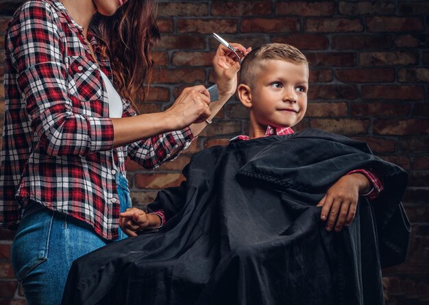 Garçon d'âge préscolaire souriant se coupe les cheveux. Le coiffeur pour enfants avec des ciseaux et un peigne coupe un petit garçon dans la chambre avec un intérieur loft.