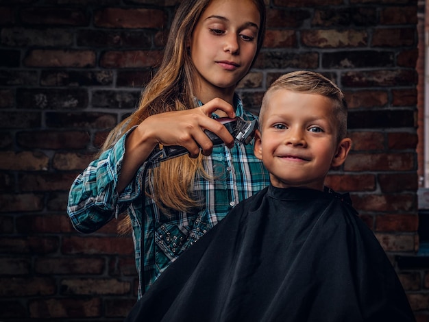 Garçon d'âge préscolaire souriant se coupe les cheveux. La sœur aînée coupe son petit frère avec une tondeuse contre un mur de briques.