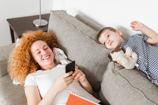 Garçon allongé sur le canapé près de sa mère heureuse à l&#39;aide de téléphone portable