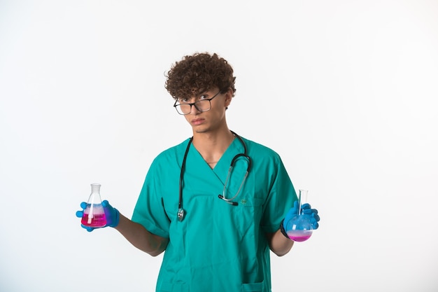 Garçon aux cheveux bouclés en uniforme médical et masques à main faisant des tests dans des flacons chimiques et avoir un visage sérieux