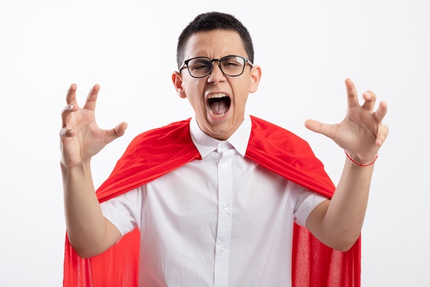 Garçon jeune super-héros en colère en cape rouge portant des lunettes en gardant les mains en l'air regardant la caméra crier isolé sur fond blanc