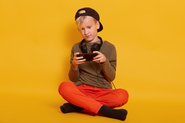 Garçon jouant au jeu via téléphone portable, adorable enfant mâle assis isolé sur jaune et tenant mobile, mec s'habille avec désinvolture, posant avec des écouteurs autour du cou, en gardant les jambes croisées.